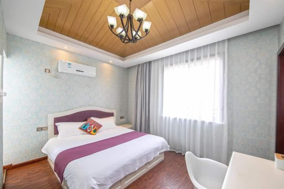 A hotel room with a bed, a white chair, a chandelier, and a window with curtains.