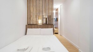 A hotel room with a bed, white linens, a wooden headboard, a bedside table with a lamp, and a television on the wall. - VLC HOST - San Jacinto A (Valencia)
