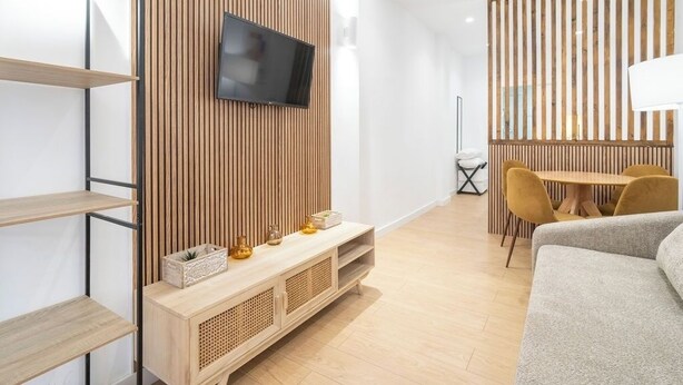 A modern living room with a wooden panel wall, a flat-screen TV, a wooden console table with decorative items, a sofa, a dining area with a table and chairs, and a floor lamp. - VLC HOST - San Jacinto A (Valencia)