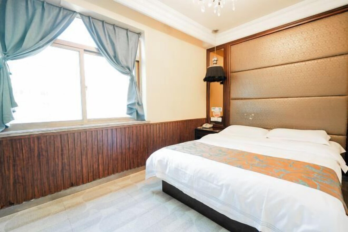 Quarto de hotel com uma cama grande, cabeceira de madeira, mesa de cabeceira, luminária e janela com cortinas.