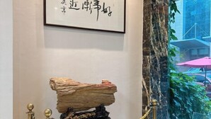 A framed calligraphy piece on a wall, a rock sculpture on a pedestal, and a glass window with a view of greenery and a pink umbrella.