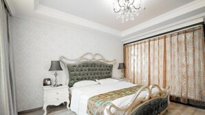 A bedroom with a tufted headboard bed, white nightstands, a chandelier, and a large window with curtains.