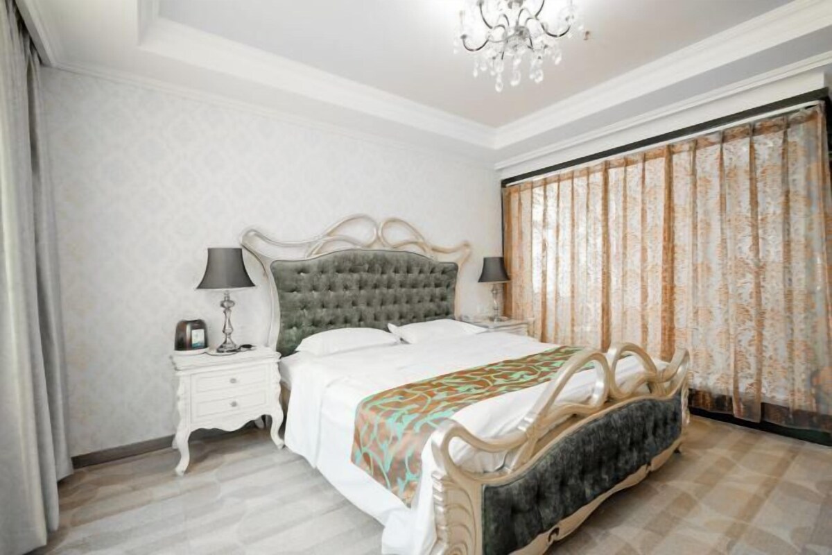 Quarto com cama de cabeceira estofada, mesas de cabeceira brancas, um candelabro e uma janela grande com cortinas.