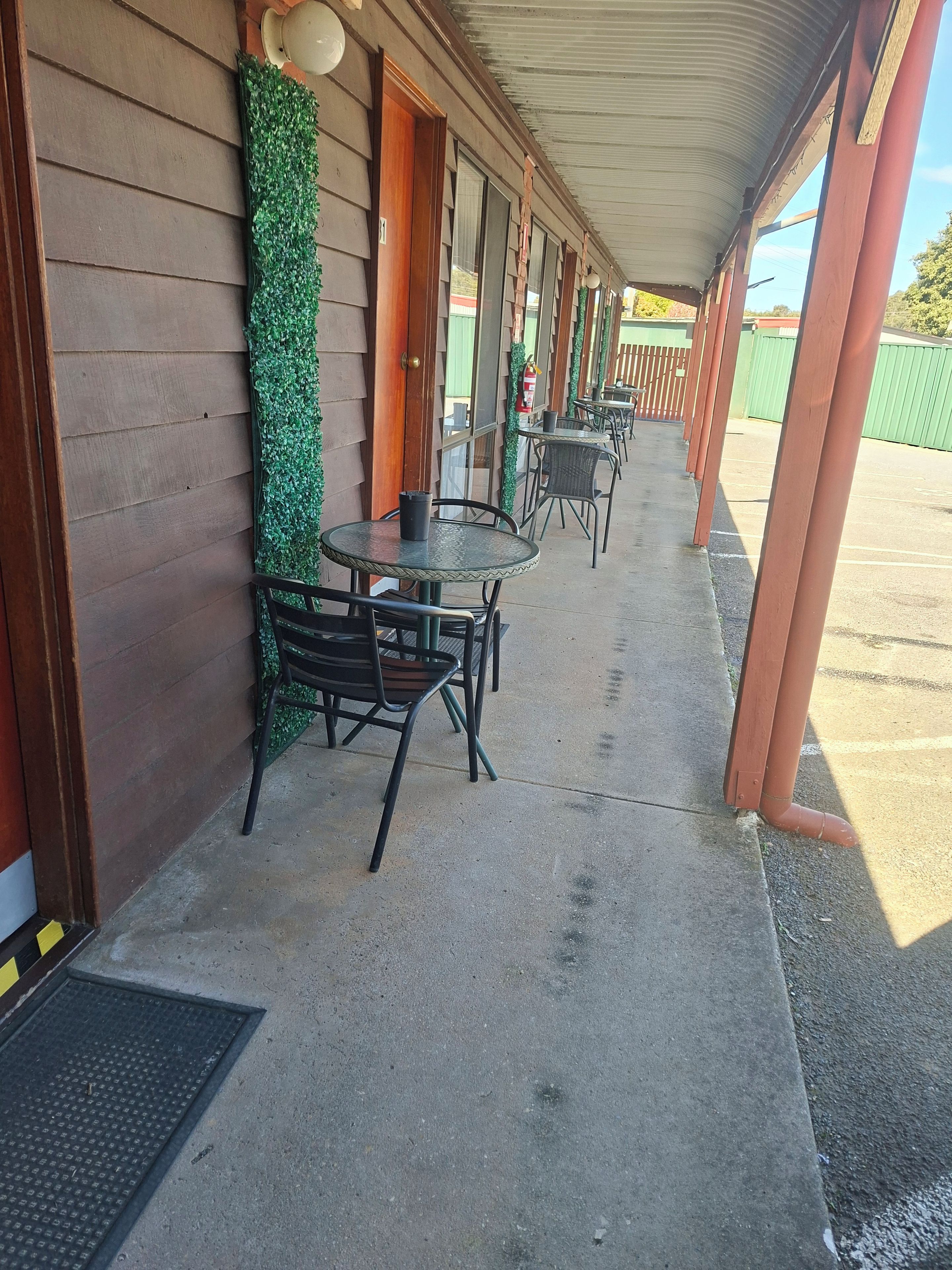 A covered outdoor seating area with tables and chairs, a fire extinguisher, and a green wall with foliage.