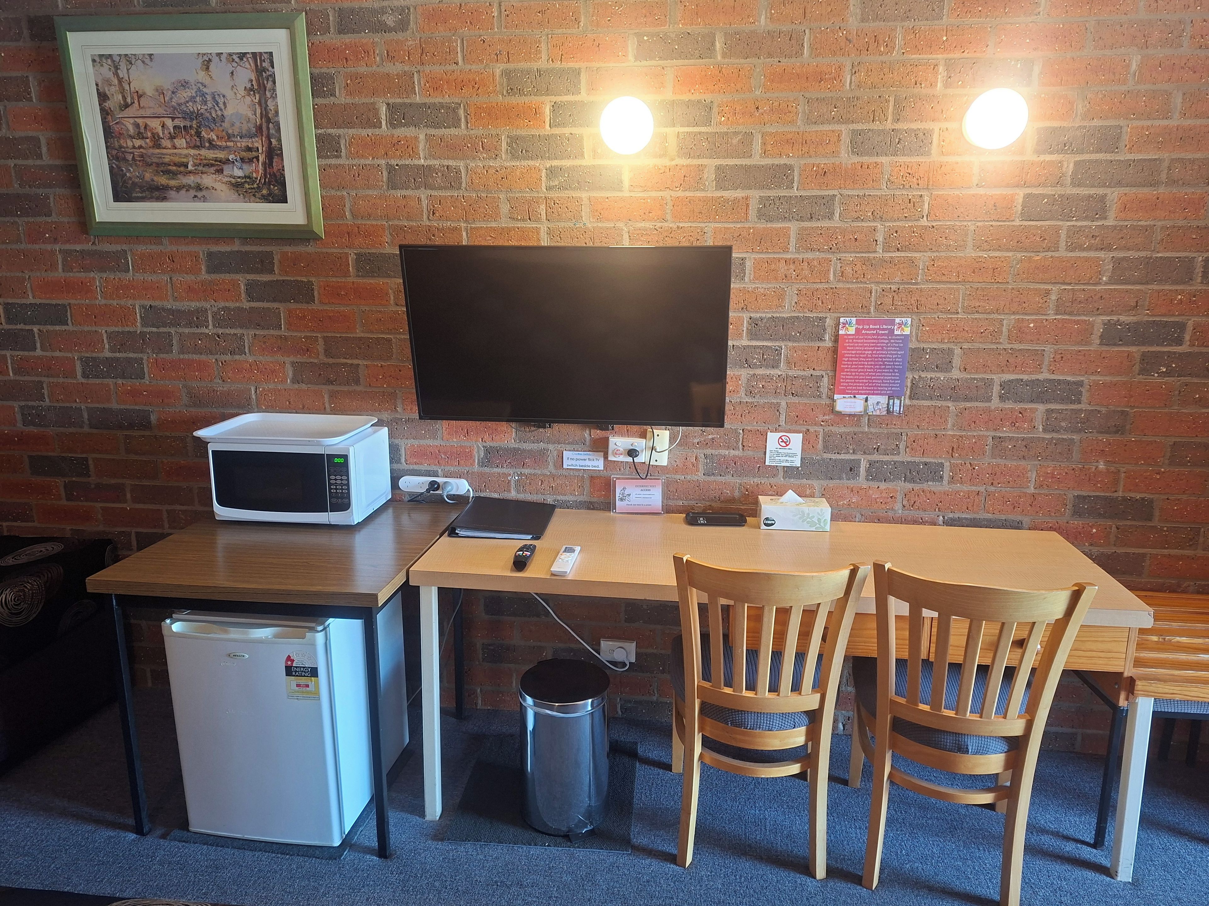A compact room with a brick wall, a flat-screen TV mounted above a wooden table, a microwave, a small refrigerator, and two wooden chairs.