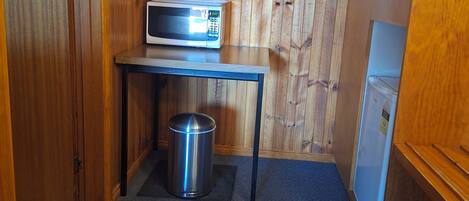 Habitación compacta con pared de madera, una mesita con microondas, un cesto de basura y un refrigerador.