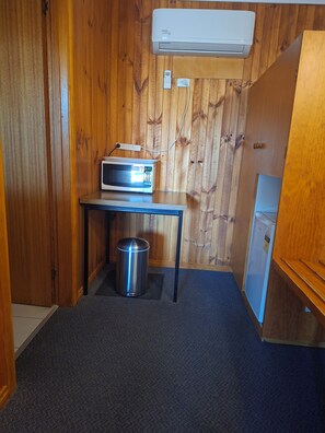 A compact room with a wooden wall, a small table with a microwave, a trash bin, and a refrigerator. - Country Road Motel (St. Arnaud)