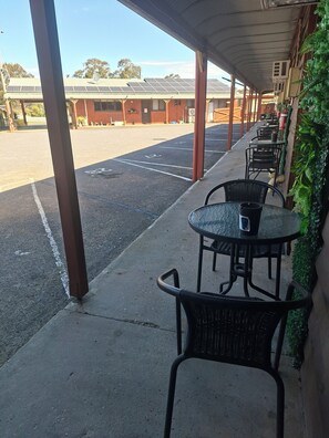 A covered outdoor seating area with tables and chairs, a parking lot in the background, and a building with a solar panel roof. - Country Road Motel (St. Arnaud)