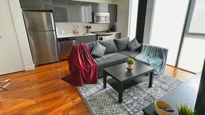 Una sala de estar moderna con un sofá gris, una mesita de centro y una planta pequeña. Se ve un área de cocina al fondo, con un microondas y un refrigerador.