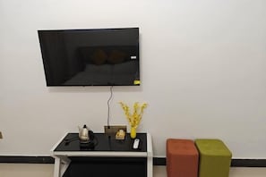 A room with a flat-screen TV mounted on the wall, a small table with a remote, a decorative vase, and two colorful cushions.