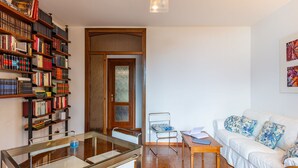 Ein modernes Wohnzimmer mit einem Glastisch, einem weißen Sofa mit gemusterten Kissen, einem Bücherregal voller Bücher und einer Tür mit Milchglasfenstern.