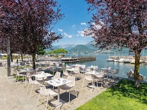 An outdoor café with tables and chairs, a body of water, boats, and mountains in the background. - Ortensie by Interhome (Colico)