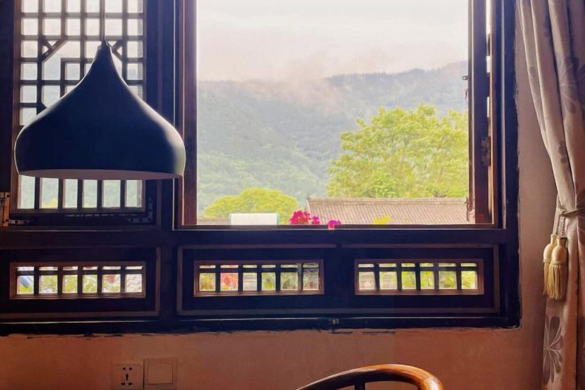 A room with a large window featuring traditional wooden lattice design, a black hanging lamp, and a view of a green landscape with hills.