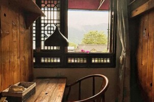 Una habitación de madera con una mesa, una silla y un ventanal con vistas a zonas verdes y montañas, con una lámpara colgante.