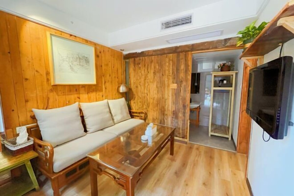 A wooden-paneled room with a sofa, a glass coffee table, a television, and a door leading to a kitchen.