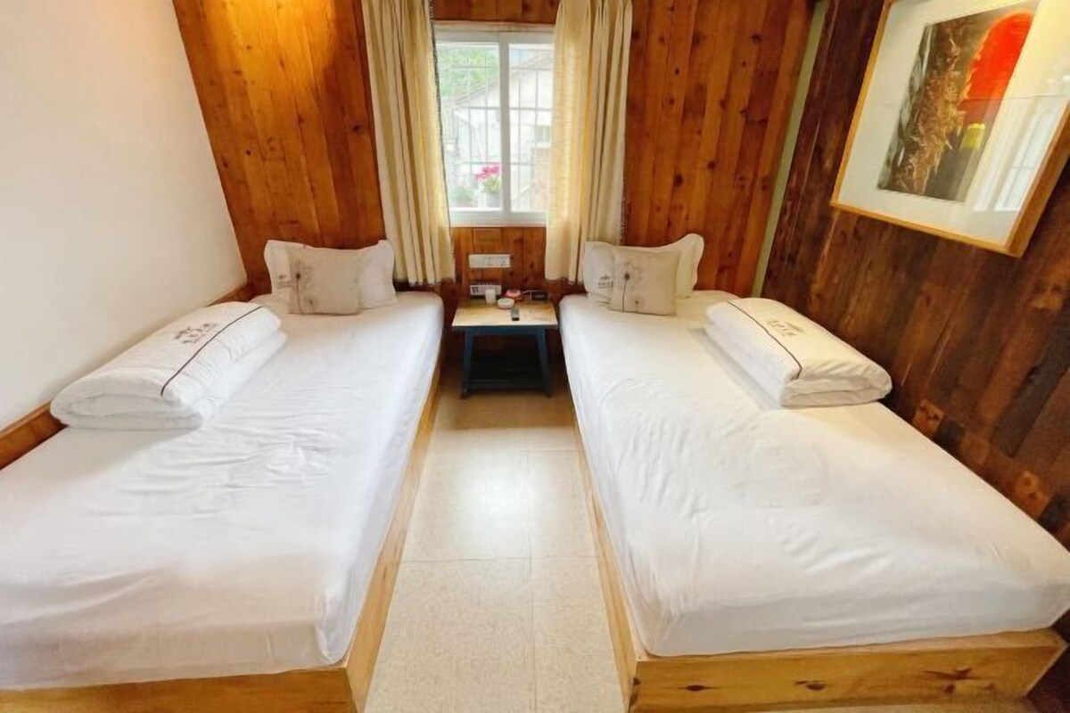 A room with two single beds, wooden walls, a small table with a lamp, and a window with curtains.