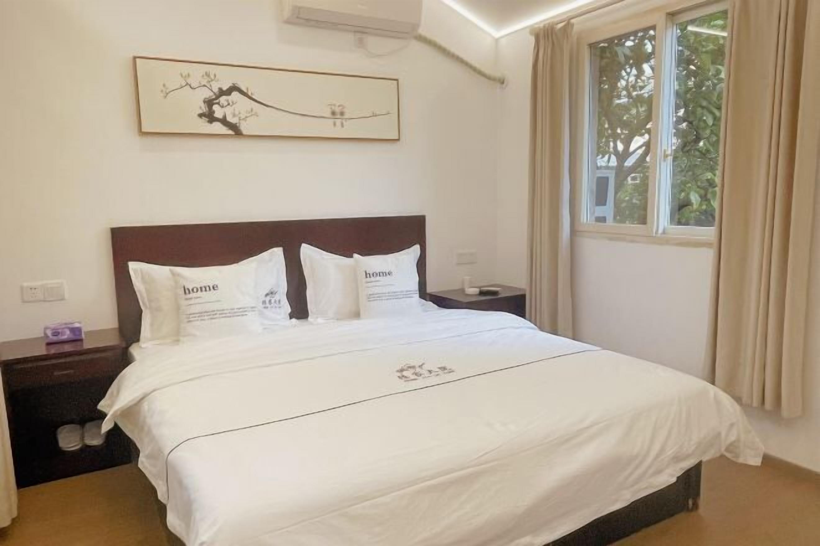 A neatly made bed with white bedding and pillows, a dark wooden headboard, a small bedside table with a lamp, and a window with curtains.