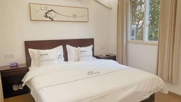 Cama bem arrumada com roupa de cama e almofadas brancas, cabeceira de madeira escura, pequena mesa de cabeceira com abajur e uma janela com cortinas.