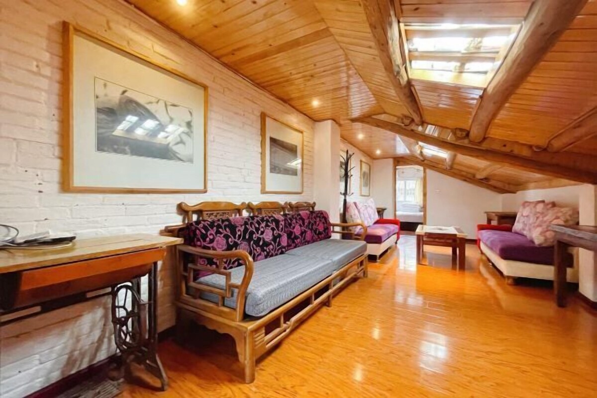 A cozy loft bedroom with a wooden ceiling, a bed with a patterned mattress, a wooden desk, and a large window providing natural light.