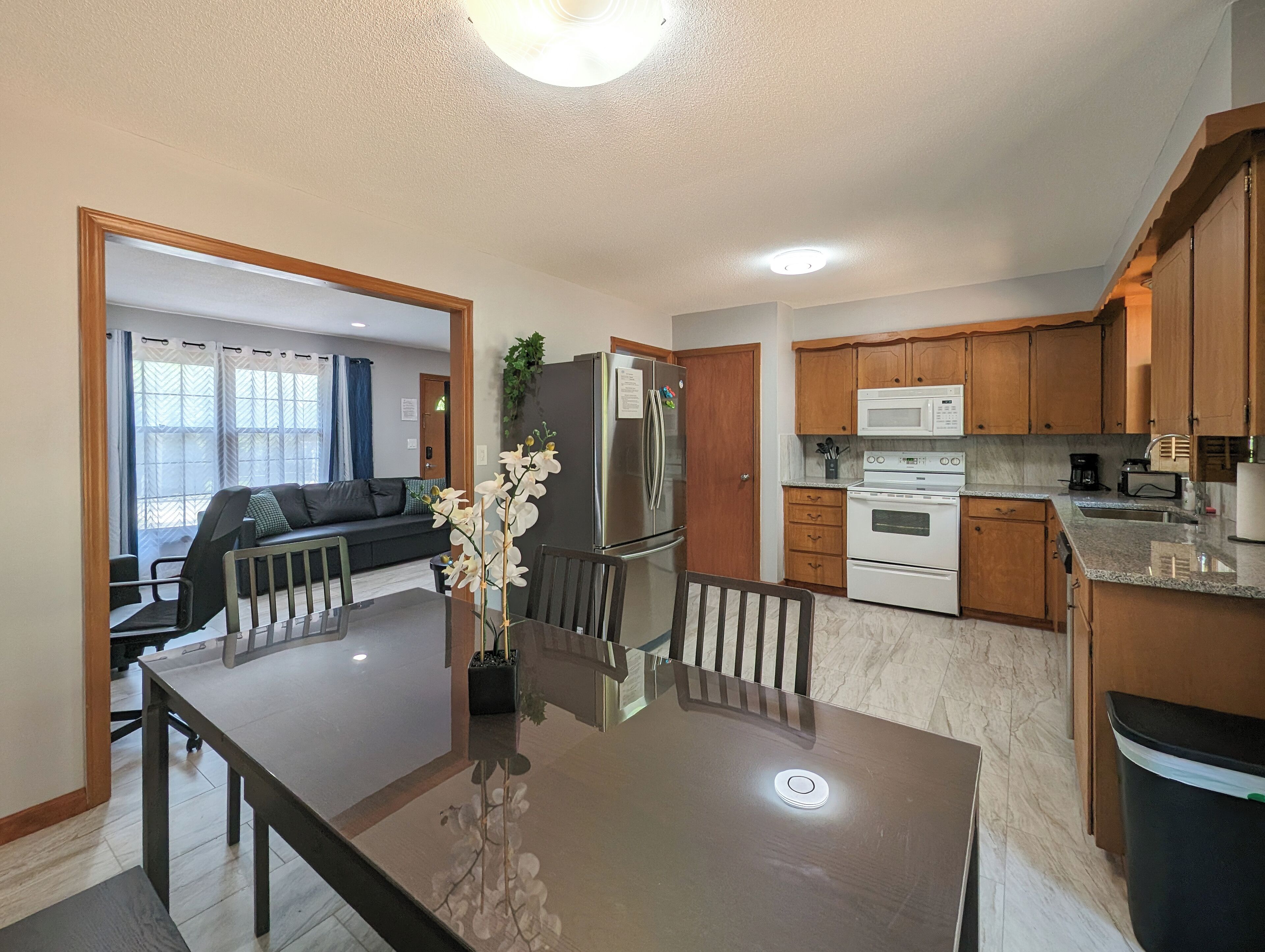 A modern kitchen with wooden cabinets, a stainless steel refrigerator, a microwave, and an oven. There is a dining table with chairs and a vase of flowers.
