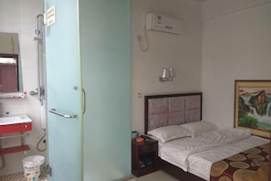 A hotel room with a bed, a nightstand, a wall-mounted air conditioner, a painting, and a bathroom with a sink and mirror.