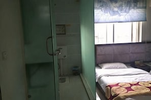 Een kamer met een bed, een raam met gordijn, een douchegedeelte en een klein tafeltje met een lamp.