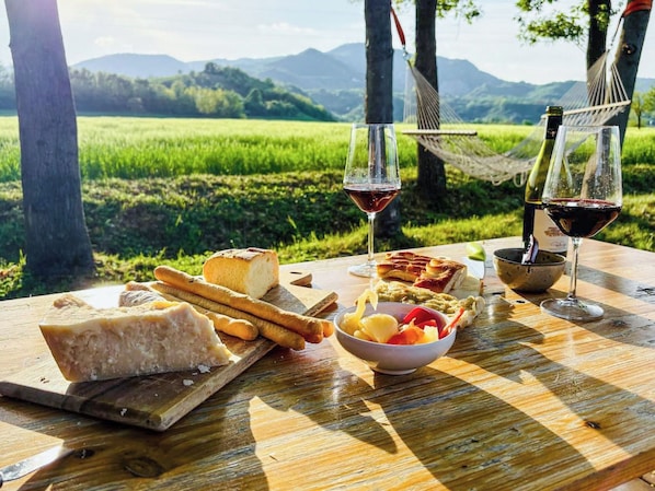 Un montaje para picnic con queso, pan y fruta sobre una mesa de madera, dos copas de vino tinto y una hamaca en un entorno natural pintoresco.