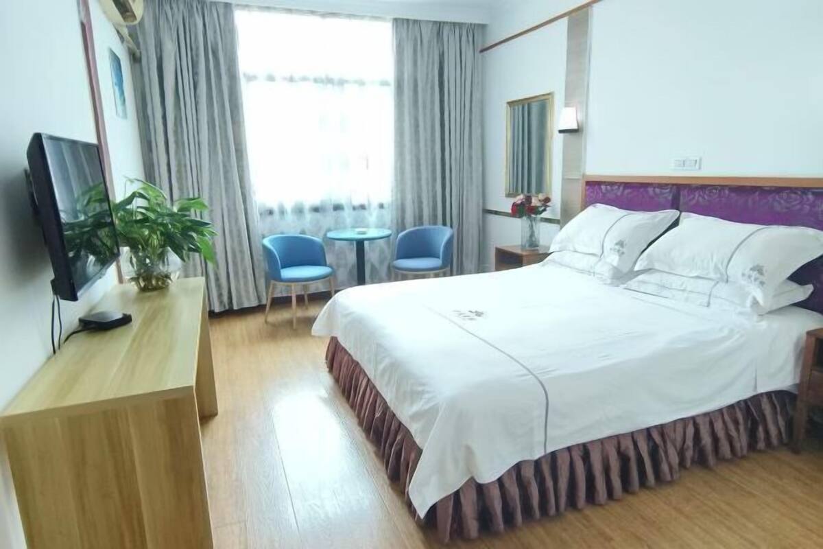 A hotel room with a bed, a desk, a TV, a plant, a chair, a table, and a window with curtains.