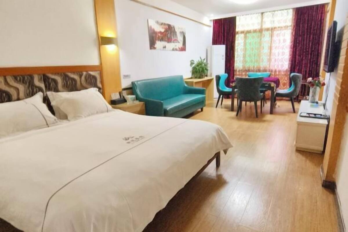 A hotel room with a large bed, a sofa, a dining table with chairs, a TV, and a plant.