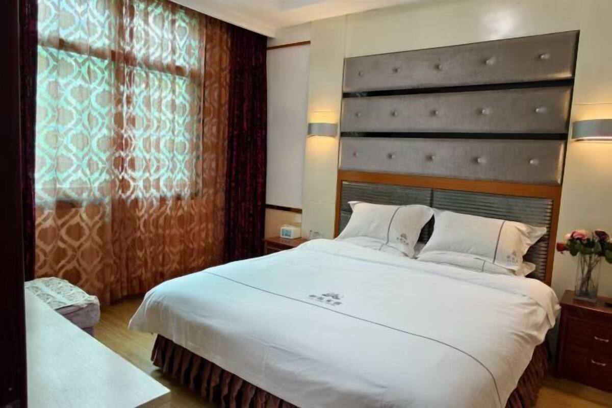 A hotel room with a large bed, a headboard with multiple sections, a bedside table, a nightstand, and a window with patterned curtains.
