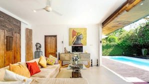A spacious living room with a large sofa, a coffee table, and a television. There is a wooden cabinet with decorative items and a painting on the wall. A pool is visible through the open space. - Villa Mangga (Legian)
