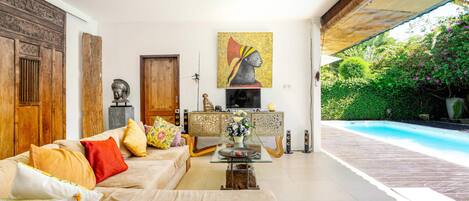 Amplia sala de estar con un sofá grande, una mesa de centro y un televisor. Se observa un mueble de madera con objetos decorativos y un cuadro en la pared. Se ve una piscina a través del espacio abierto.
