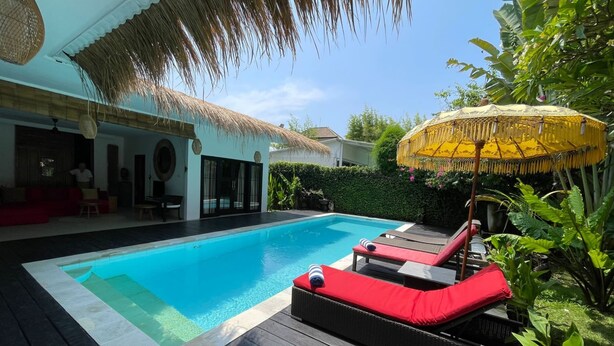 A swimming pool with lounge chairs and an umbrella, surrounded by greenery and a thatched roof structure. - Villa Mangga (Legian)