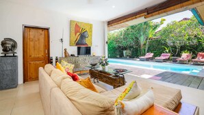 A spacious living area with a beige sofa, a wooden coffee table, and a painting of a pharaoh's head. A pool is visible through the open space. - Villa Mangga (Legian)