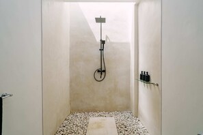 Un petit espace douche fermé, doté d’une seule pomme de douche, d’une étagère en verre pour les articles de toilette et d’un sol recouvert de petits galets.