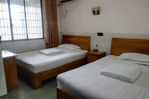 Habitación de hotel con dos camas individuales, cabecera de madera, mesita de noche y persianas en la ventana. Cuenta con aire acondicionado instalado en la pared.