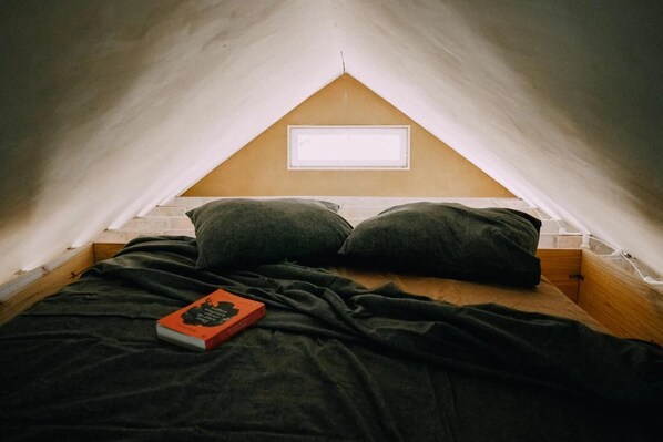 Une petite chambre mansardée de forme triangulaire, avec un lit simple, deux oreillers et un livre posé sur le lit.
