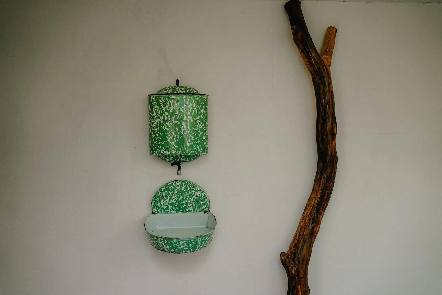 Un récipient mural vert et blanc à motifs, doté d’un couvercle et d’un petit bol placé en dessous, le tout accompagné d’une branche en bois.