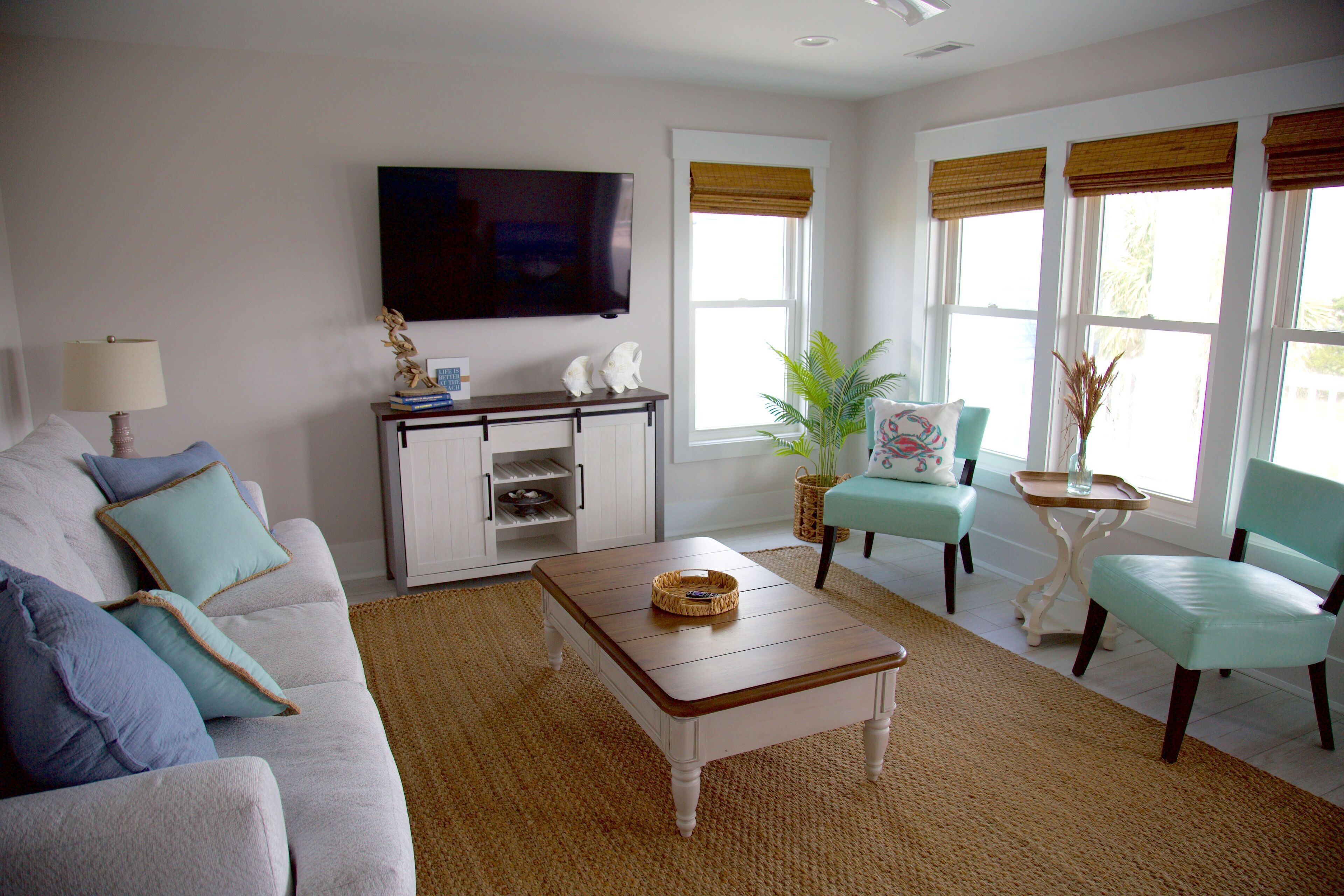 A living room with a sofa, coffee table, and two chairs. There is a television on the wall and a window with blinds.