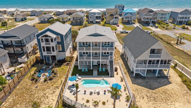 A coastal residential area with multiple two-story houses, a central swimming pool, and a beach in the background. - New!  Spectacular Ocean & Marsh Views, Elevator, Pool, Den, ADA, Steps to Beach! (Oak Island)