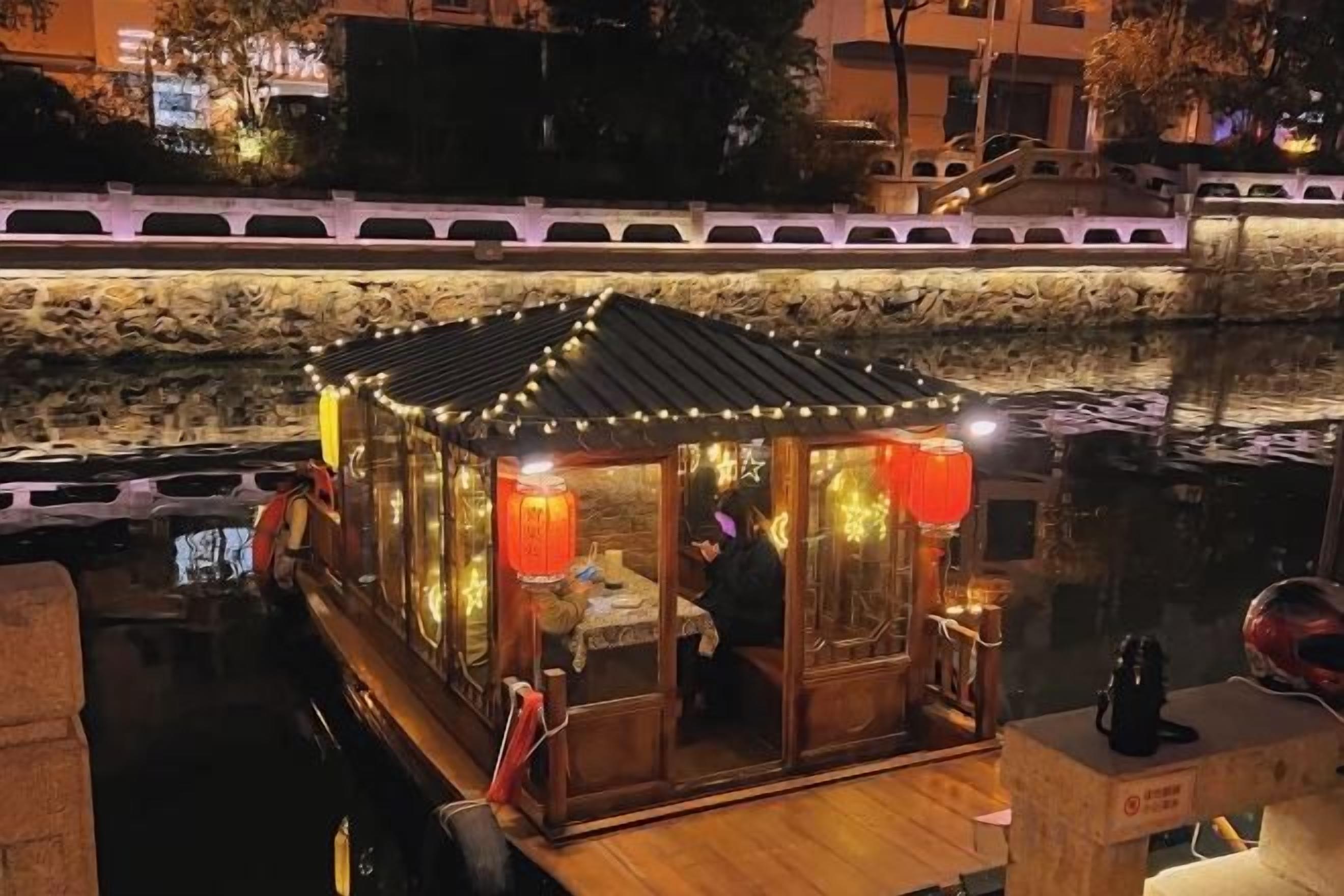 A traditional wooden boat with a roof, illuminated by string lights, docked at a riverside.