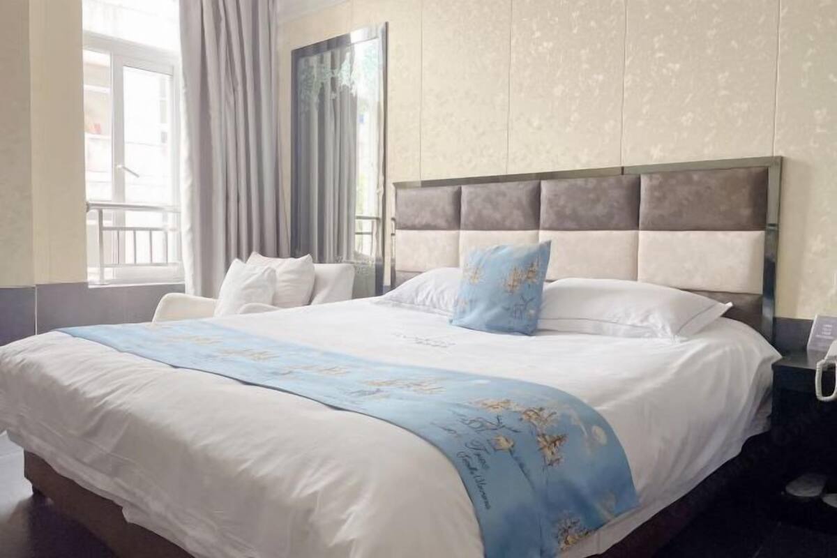 A hotel room with a large bed, a headboard, a bedside table with a phone, and a window with curtains.