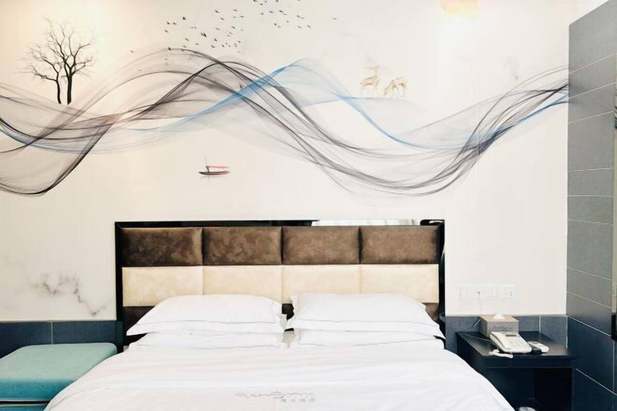 A modern hotel room with a large bed, a headboard with a padded design, a bedside table with a phone, and a wall with abstract art.