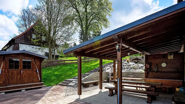 A rustic wooden building with a covered patio area, outdoor seating, and a stone staircase leading up to a higher level.