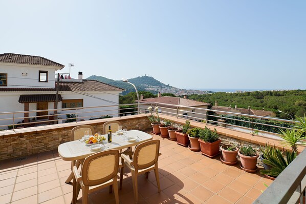 A terrace with a table set for two, surrounded by potted plants, overlooking a scenic landscape with buildings and hills. - 2MARINA01 - House with a private pool in a residencial area (Blanes)