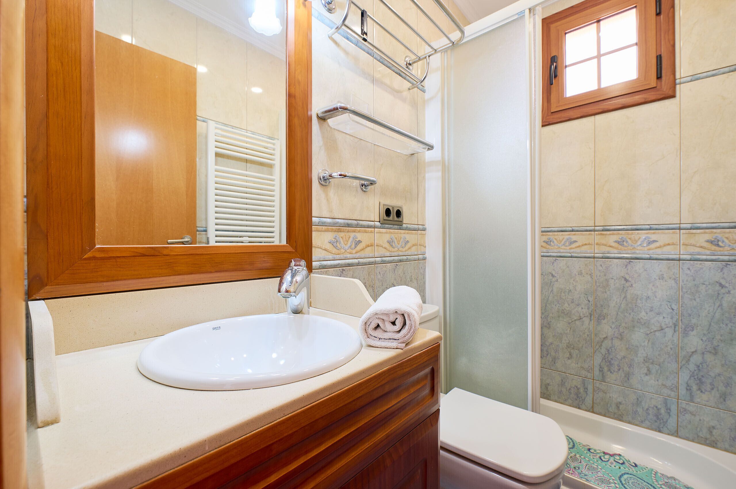 Un baño con tocador de madera, un lavabo blanco, una toalla, ducha con puerta de vidrio y una ventana.