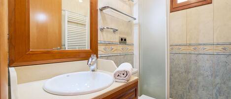 Un baño con tocador de madera, un lavabo blanco, una toalla, ducha con puerta de vidrio y una ventana.