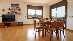 A spacious living area with a flat-screen TV, wooden entertainment unit, dining table with chairs, and large windows offering a view of the sea and hills.