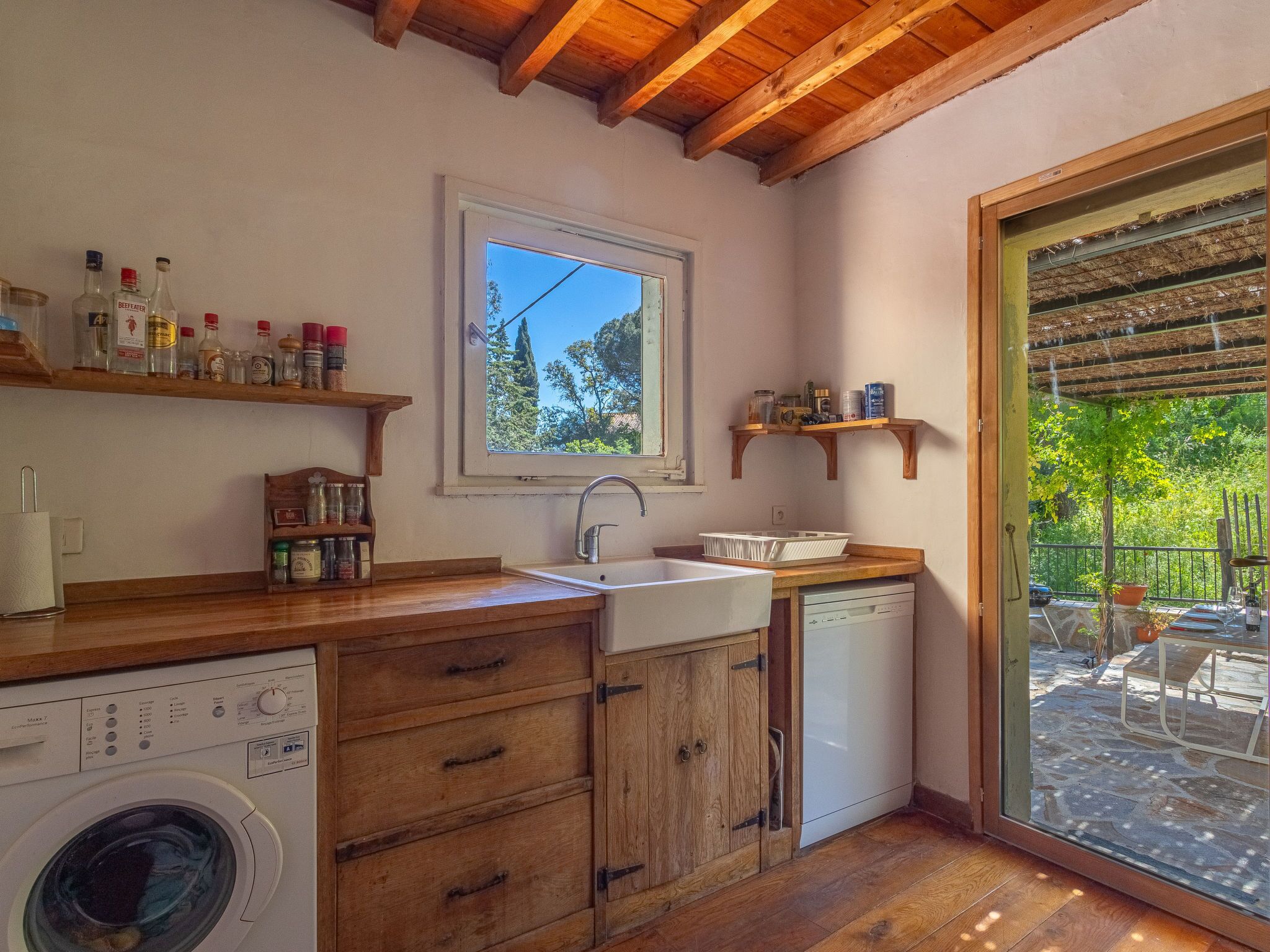 A compact kitchen with wooden cabinets, a washing machine, a sink, and a window overlooking greenery.