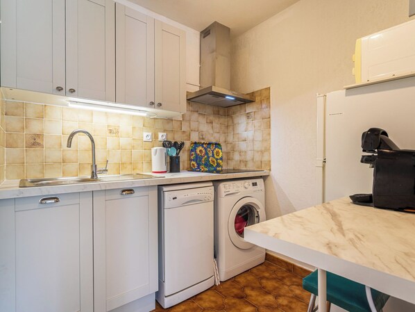 A compact kitchen with white cabinets, a built-in dishwasher, a washing machine, and a countertop with a coffee machine.